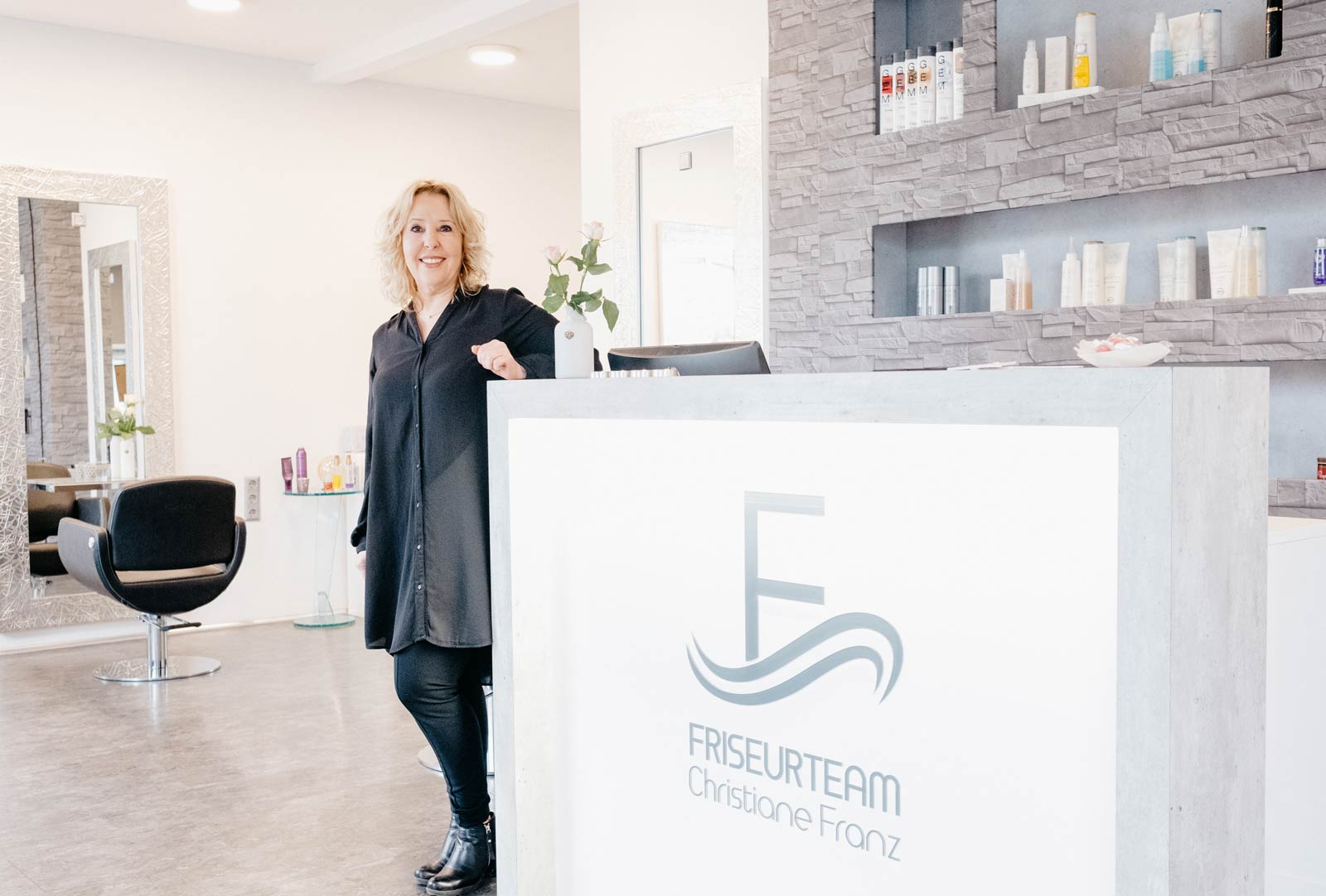 Christiane Franz lehnt lächelnd an ihrem Empfangstresen mit dem Friseurteam-Logo